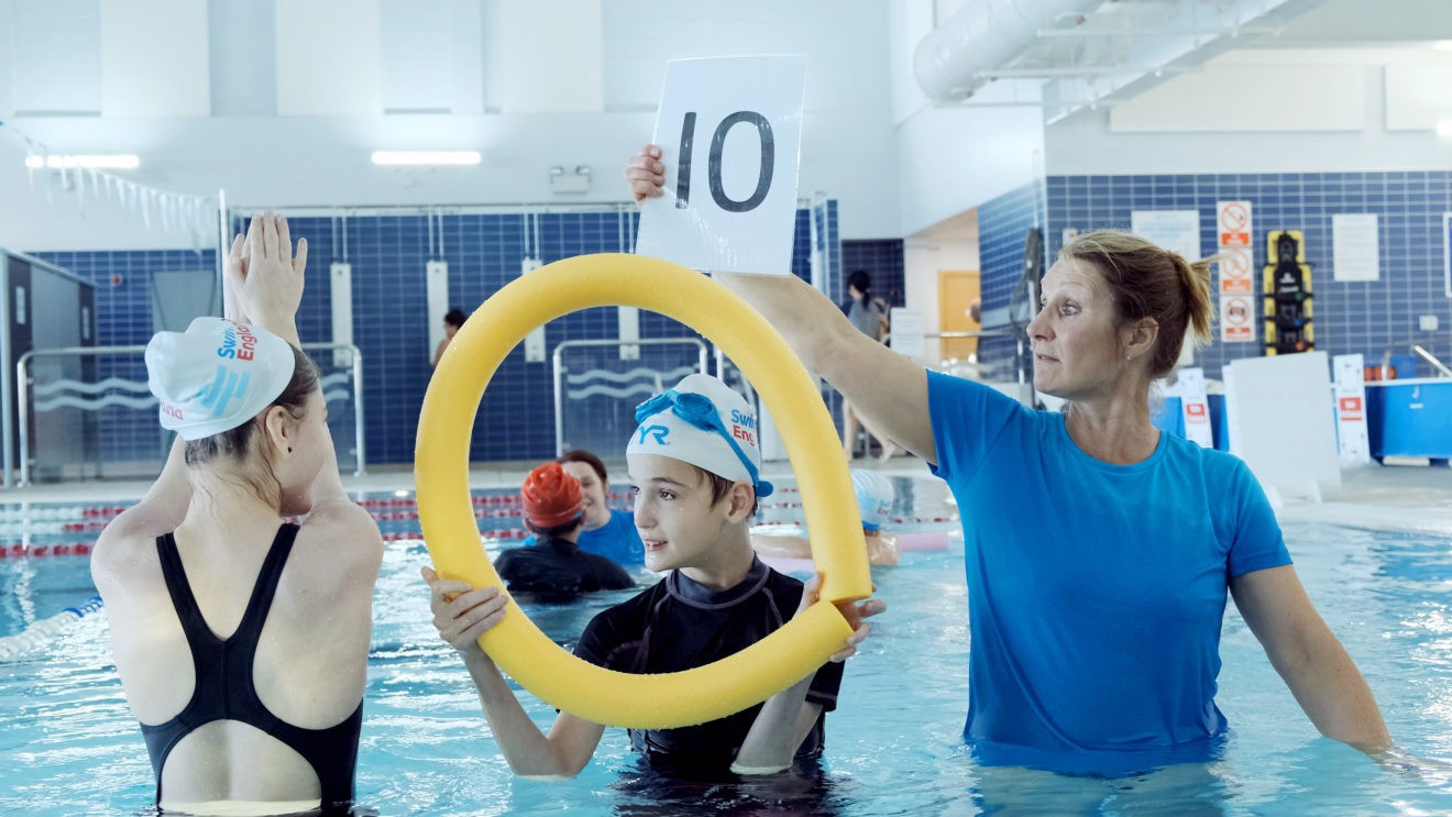Teacher holding a number 10 sign and students in swimming pool holding pool noodle